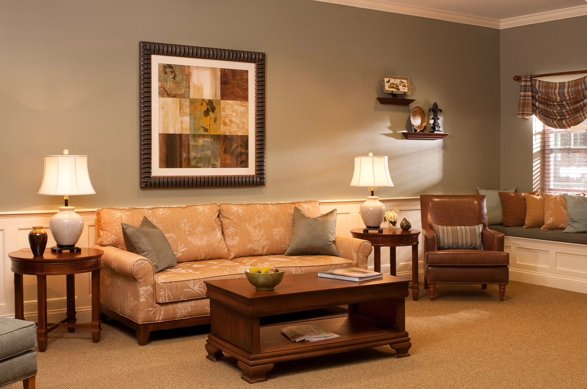 A cozy living room with a beige patterned sofa, two side tables each with a lamp, a wooden coffee table with a bowl of fruit and a book, a brown leather armchair with a striped pillow, and a window seat with multiple cushions. The walls are painted light beige with white wainscoting, and there is a framed abstract artwork above the sofa and two small shelves with decorative items on the wall.