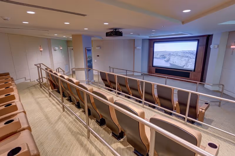 Interior view of a small theater room with two rows of cushioned seats facing a large screen displaying a nature scene. The room has beige walls, carpeted floor, and metal handrails along the aisles.