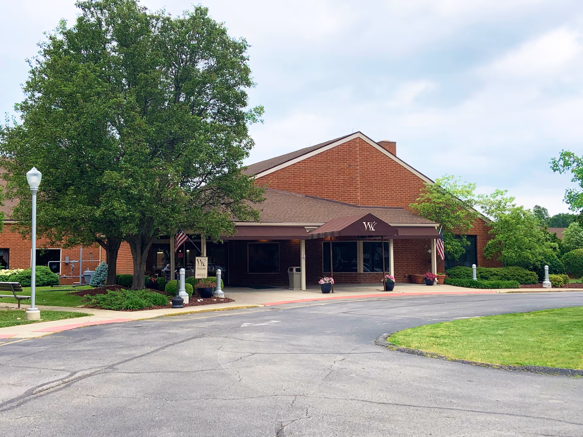 Front exterior view of Westminster Health Care Center, a single-story brick building with a covered entrance. There are trees, shrubs, and a lawn surrounding the driveway leading to the entrance. Two American flags are displayed near the entrance.