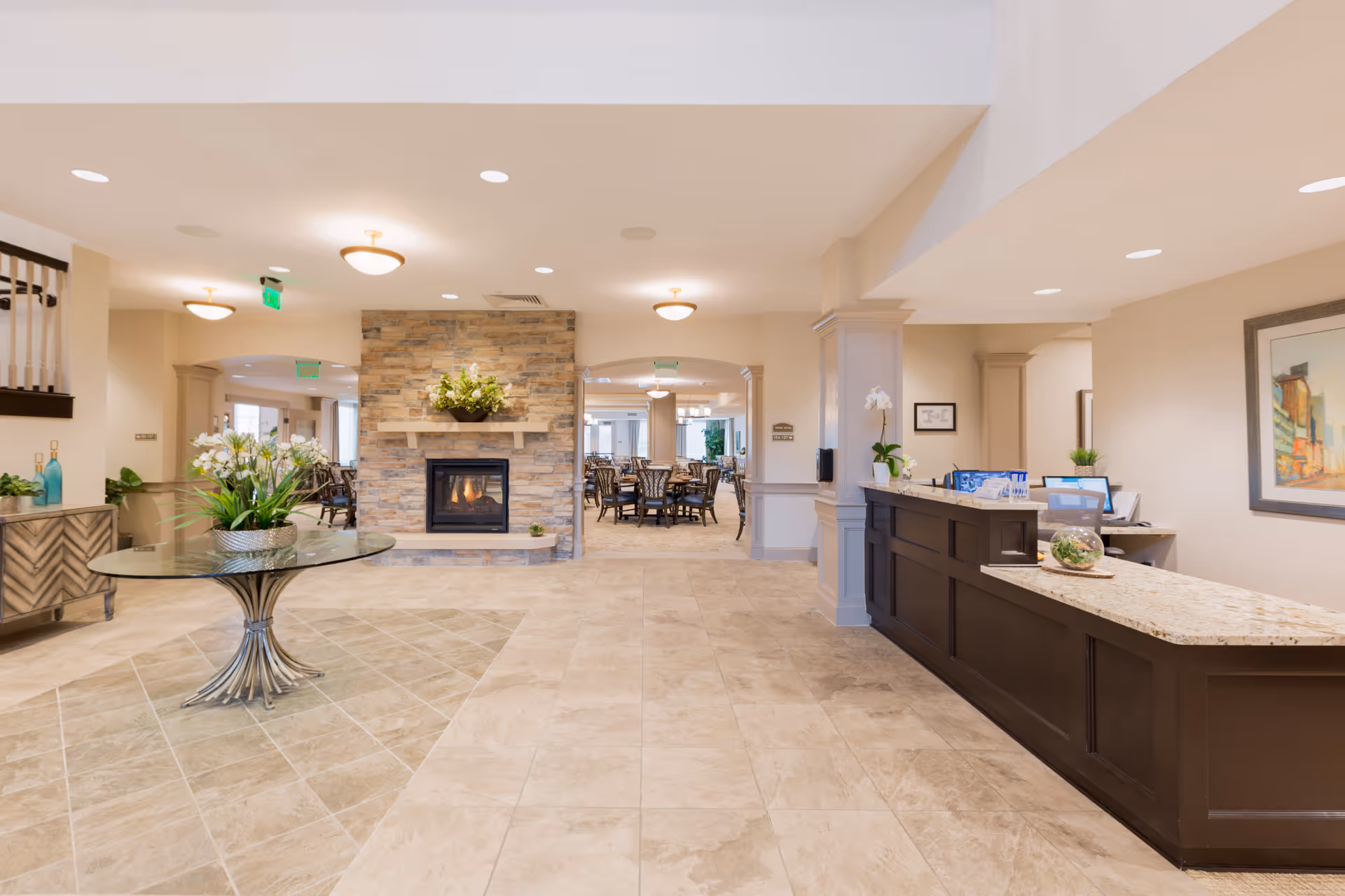 Spacious and well-lit senior living facility interior featuring a reception desk on the right, a round glass table with a floral arrangement in the center, and a stone fireplace with a flower arrangement above it. In the background, there is a dining area with multiple tables and chairs. The floor is tiled, and the walls are painted in neutral tones with recessed lighting overhead.