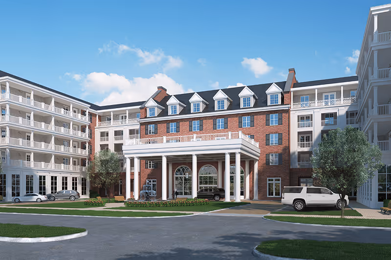 Exterior view of a large senior living facility building with a brick facade and white columns at the entrance. The building has multiple floors with balconies and windows, surrounded by a driveway with parked cars and landscaped greenery under a blue sky with some clouds.