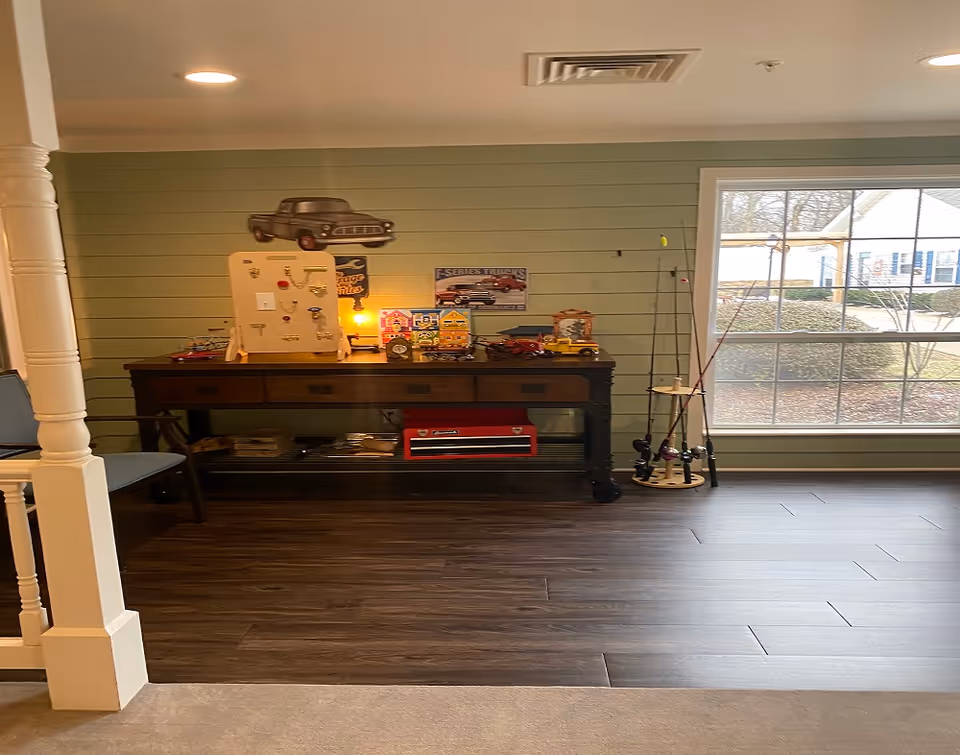 Interior room with a wooden console table against a green shiplap wall. The table holds various decorative items including toy cars, a small lamp, and a display board with keys and switches. Fishing rods are standing in a rack near a large window showing an outdoor view. A white pillar and a chair are partially visible on the left side.