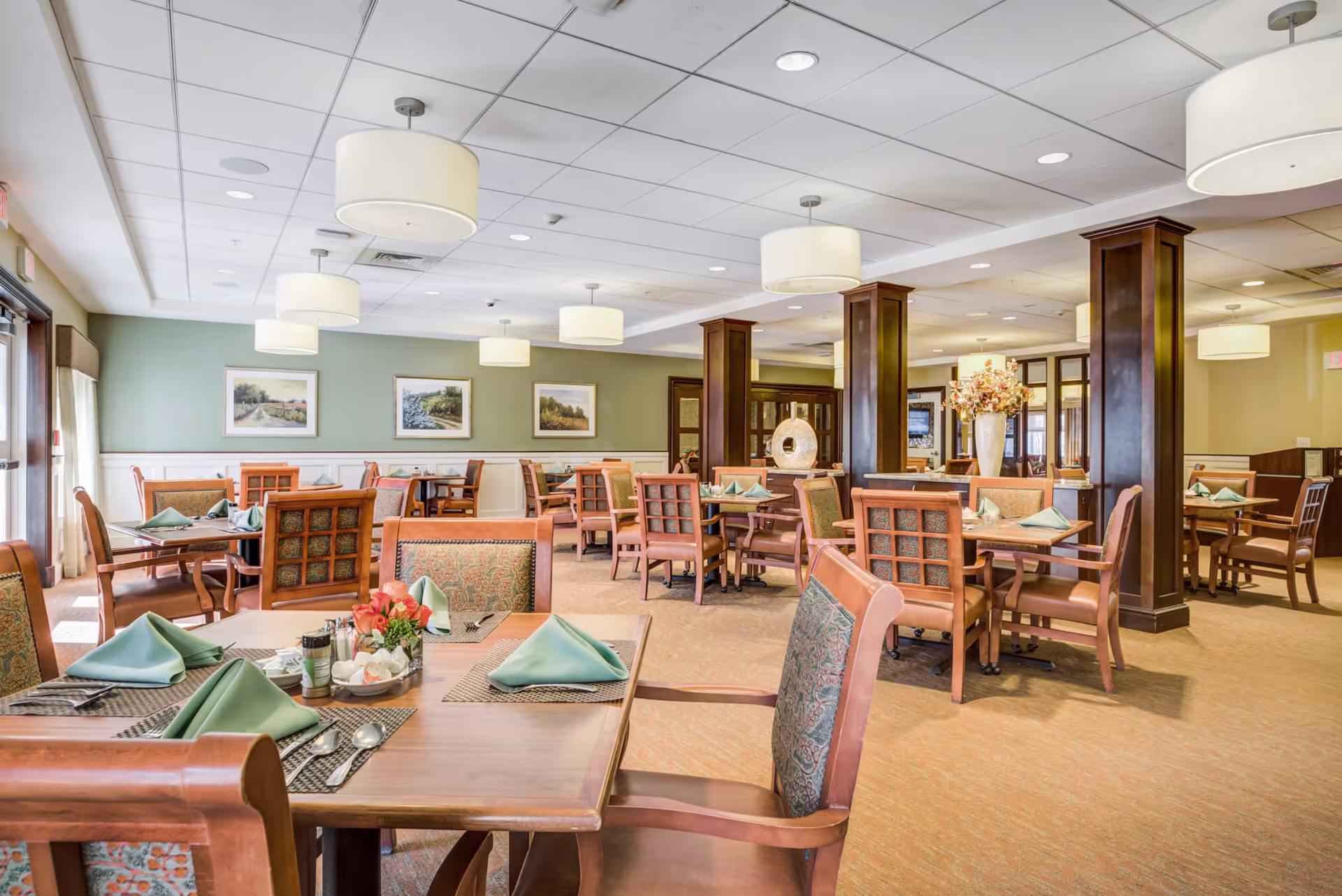 A spacious and well-lit dining room in a senior living facility with multiple wooden tables and chairs arranged neatly. Each table is set with green folded napkins, placemats, and silverware. The room features soft green walls adorned with framed landscape paintings, large ceiling lights, and wooden columns. A vase with flowers is placed on a counter near the center of the room.