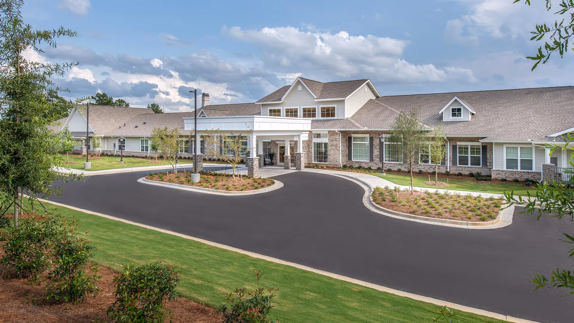 Exterior view of a single-story senior living facility building with a covered entrance, surrounded by landscaped greenery and a paved driveway under a partly cloudy sky.