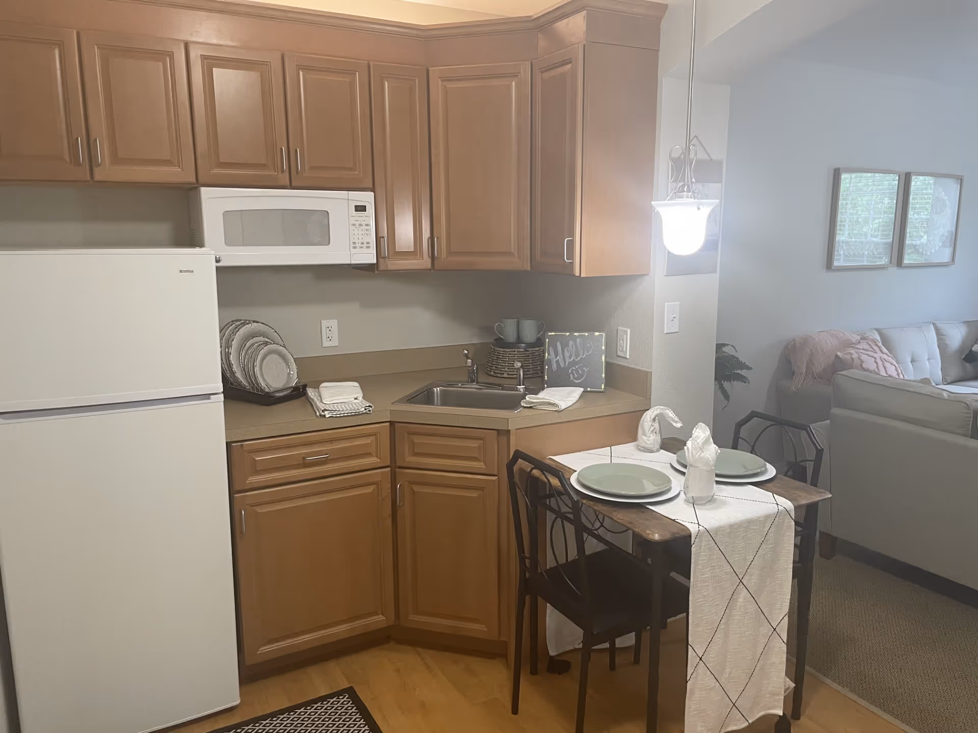 A small kitchen area with wooden cabinets, a white refrigerator, and a white microwave above the countertop. The countertop has a sink, stacked plates, and a small decorative sign that says 'Hello'. In front of the kitchen is a small dining table set for two with plates, napkins, and glasses. The background shows part of a living room with a couch and framed pictures on the wall.