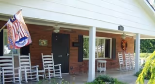 Covered porch area of a brick building with white pillars, several white rocking chairs, a small table, and an American flag hanging on the left side.