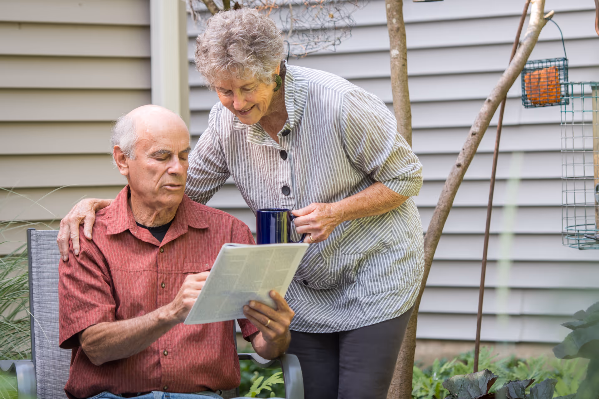 An elderly man sitting outdoors reading a newspaper while an elderly woman stands beside him holding a blue mug and smiling, with a house siding and garden in the background.
