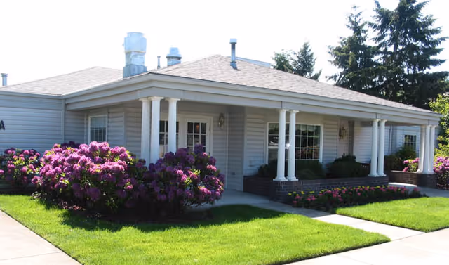 Exterior view of a single-story senior living facility building with white siding, a covered porch supported by white columns, and well-maintained landscaping including green grass and blooming purple flowers.