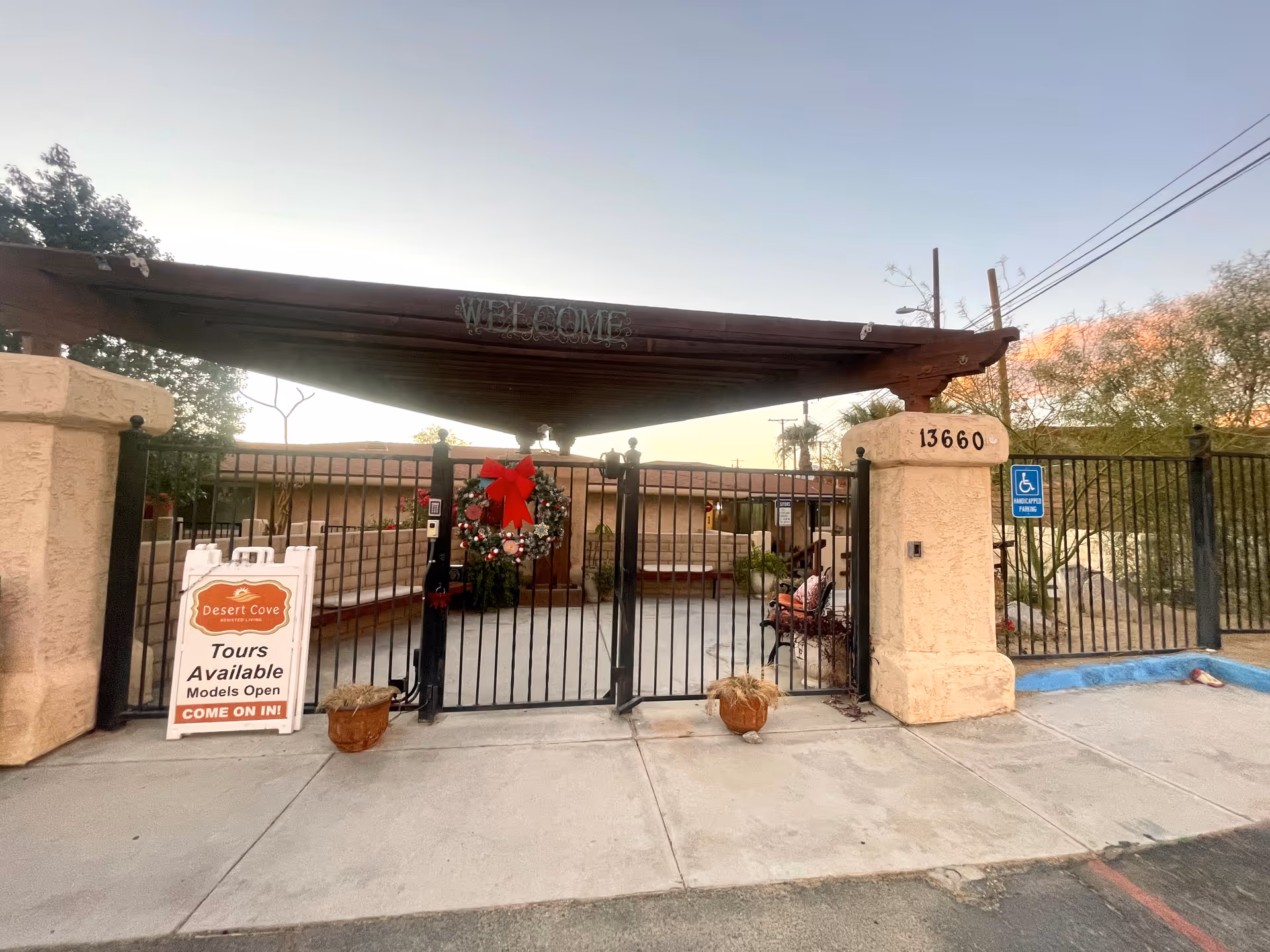 Gated front entrance of an assisted living facility with a pergola 'WELCOME' sign, holiday wreath on the gate, address number 13660, and a 'Tours Available' sign.