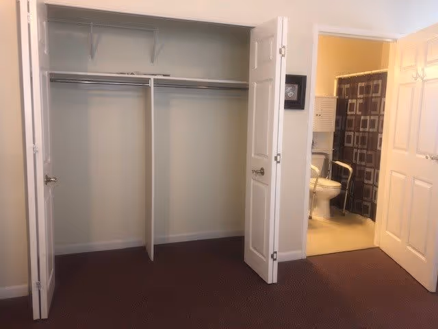 An empty closet with double white bi-fold doors open, showing two hanging rods and a shelf above. To the right, an open door reveals a bathroom with a toilet, a shower curtain with a geometric pattern, and a grab bar next to the toilet.