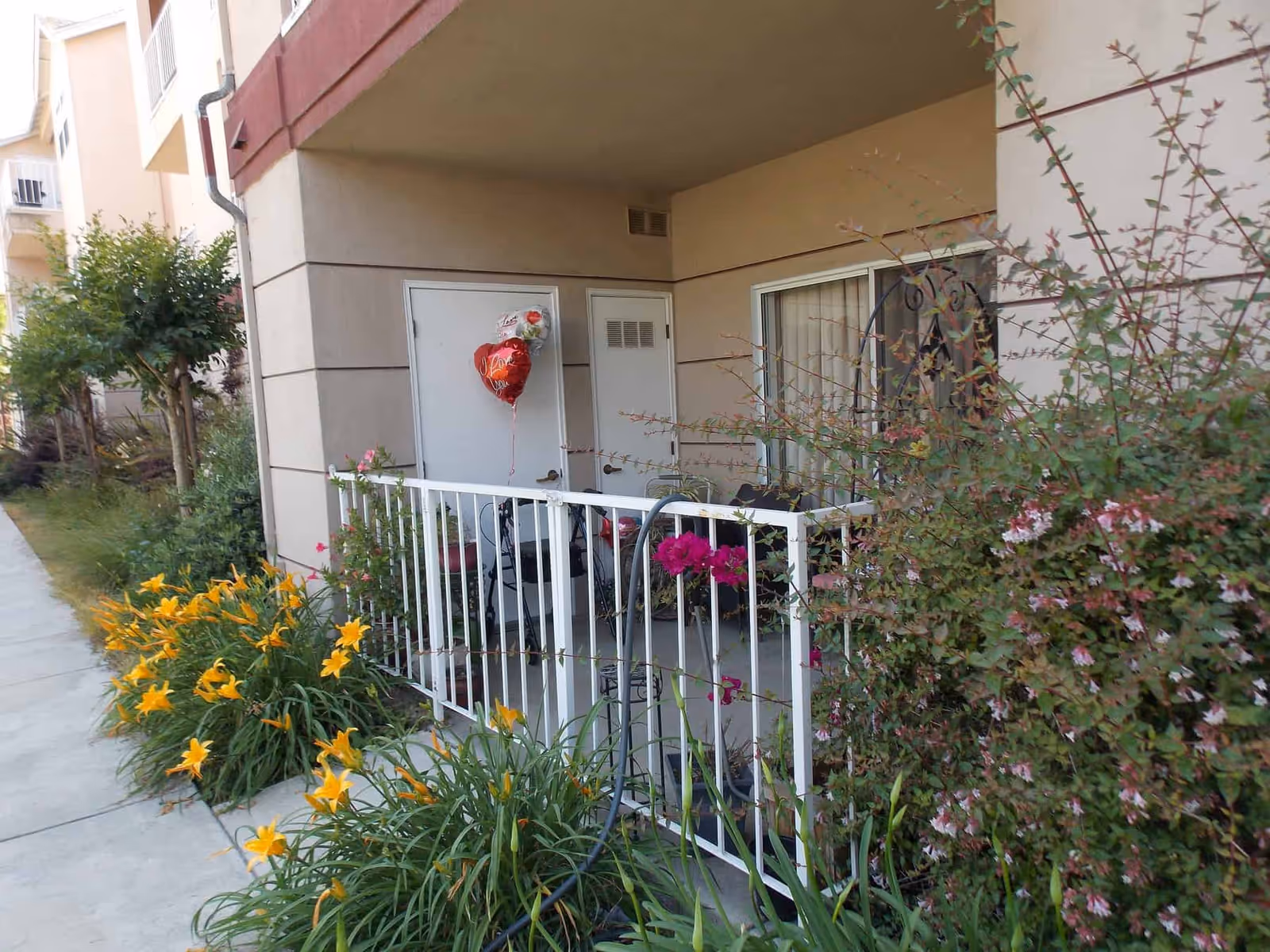 Ground-floor apartment patio with white railing, chairs, a heart-shaped 'Love' balloon, and surrounding flowering plants.