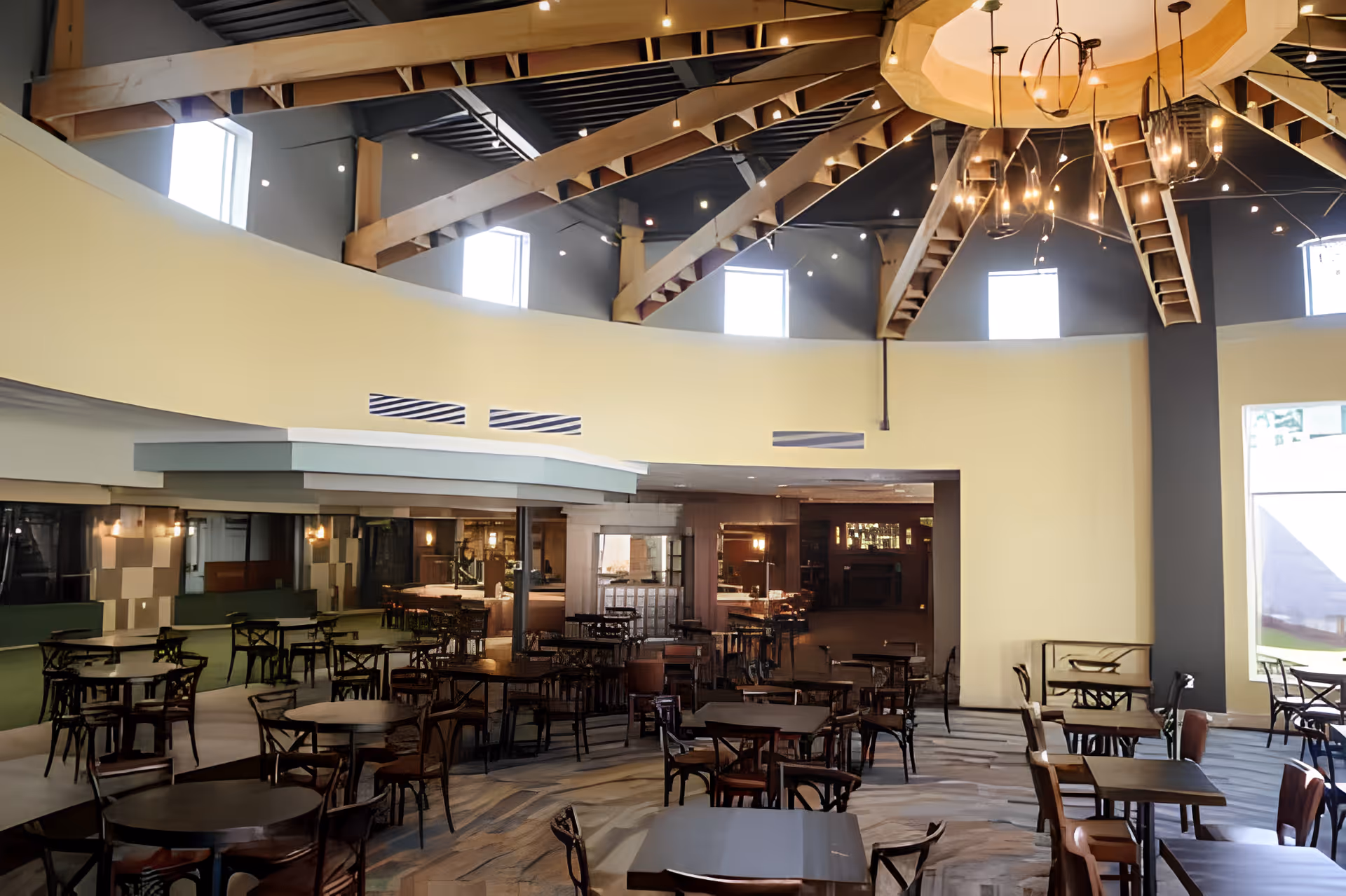 A spacious dining area with multiple dark wooden tables and chairs arranged neatly. The room features a high ceiling with exposed wooden beams and several small windows near the ceiling allowing natural light to enter. A large, modern chandelier hangs from the center of the ceiling. The walls are painted in light colors, and there is a large window on the right side letting in additional daylight.