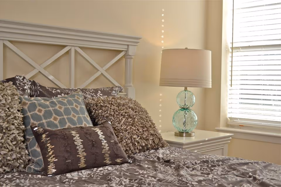 Sunlit bedroom with a white headboard, decorative pillows on a patterned bedspread and a bedside table with a glass lamp by a window with blinds.