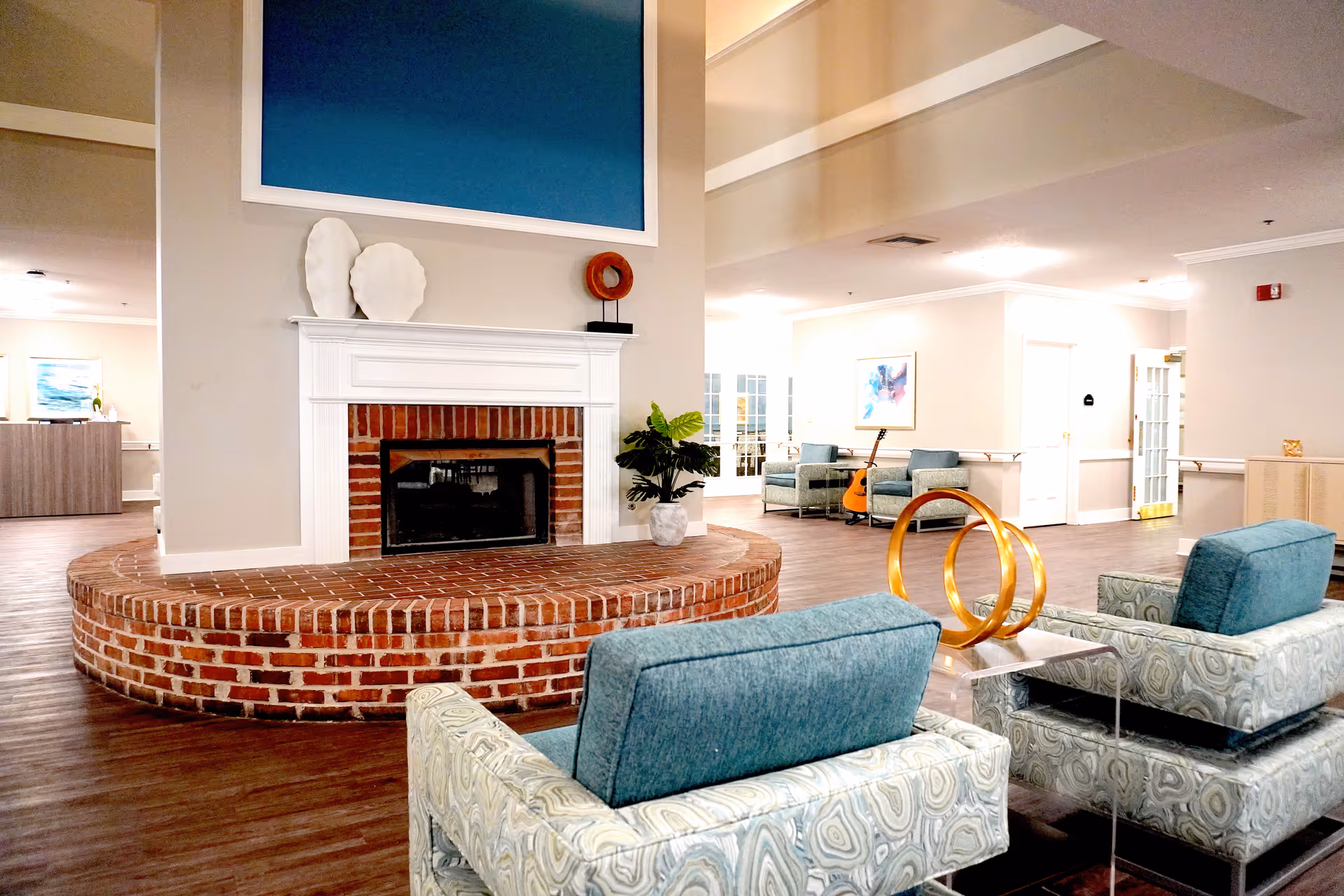 Bright senior living common area with a brick fireplace, upholstered chairs, decorative sculptures and a guitar in the background.
