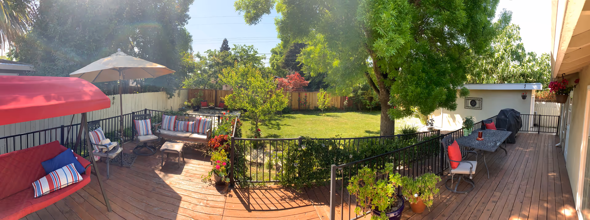 A spacious outdoor wooden deck area with seating including cushioned chairs, a bench swing with red canopy, and a table with chairs. The deck overlooks a green lawn with trees and plants, surrounded by a wooden fence. There is a large umbrella providing shade over part of the seating area and a barbecue grill near the table.