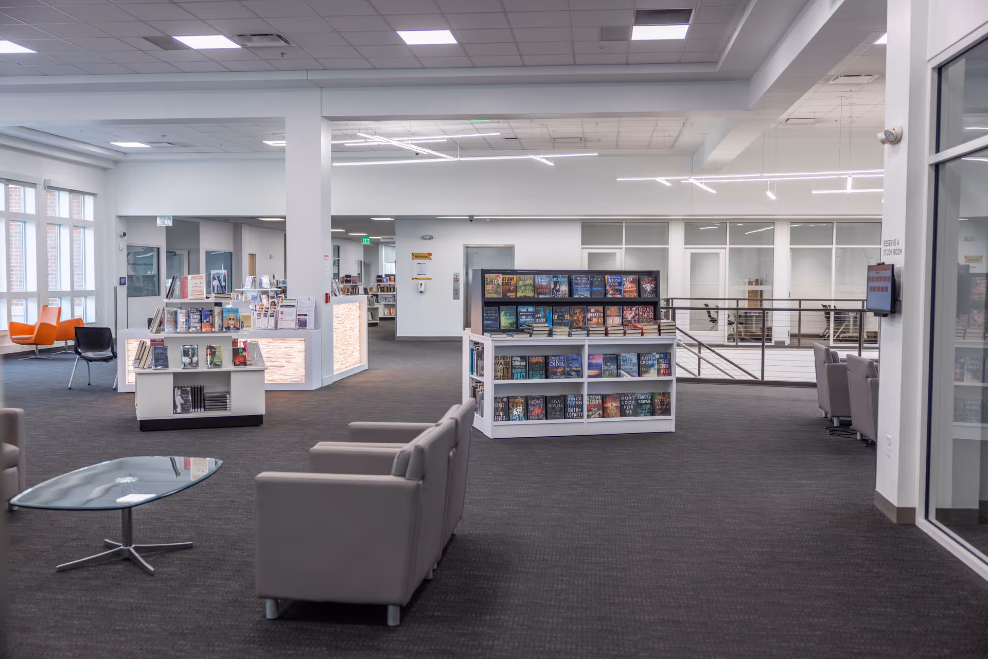 A spacious, modern library or reading area with bookshelves filled with books, several comfortable armchairs, a glass coffee table, and large windows letting in natural light. The space has a clean, open design with white walls and ceiling lights.