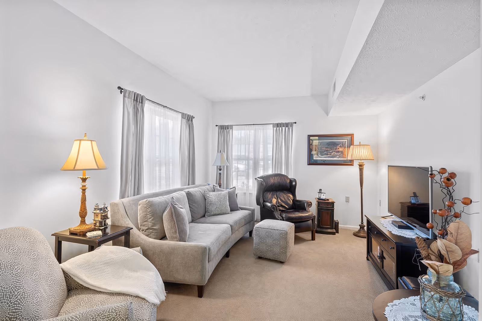 A cozy living room with beige carpet and white walls featuring a gray sofa with multiple cushions, a patterned armchair with a white throw blanket, a black leather armchair, and a small patterned ottoman. There are two windows with sheer white curtains and gray drapes. The room is lit by three lamps: a table lamp on a side table, a floor lamp near the black armchair, and another table lamp on a TV stand. The TV stand holds a flat-screen TV and decorative items including a vase with dried flowers. A framed picture hangs on the wall behind the black armchair.