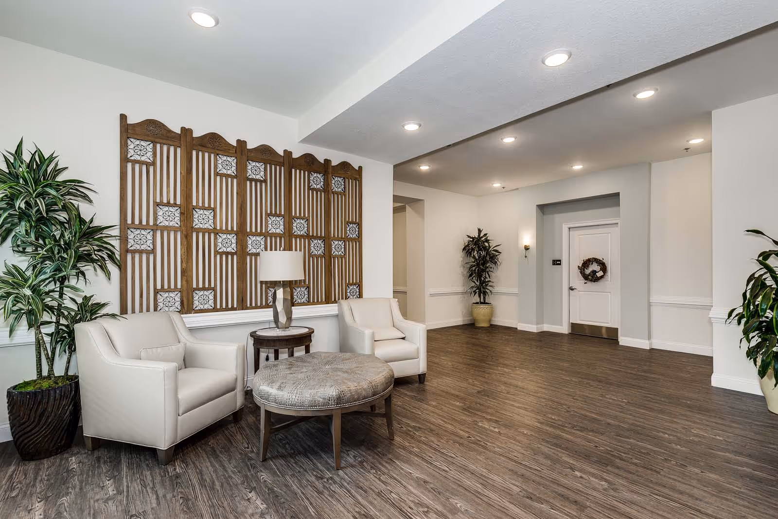 A modern and spacious interior lounge area with two white armchairs, a round ottoman, a small side table with a lamp, decorative wooden wall art, and several potted plants. The room has wood flooring, white walls, recessed ceiling lights, and a white door with a wreath.
