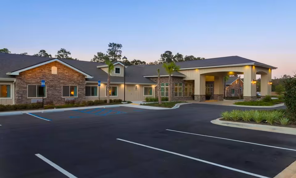 Exterior view of Azalea Gardens Assisted Living & Memory Care building at dusk, showing a single-story structure with stone and siding facade, a covered entrance, palm trees, and an empty parking lot with marked spaces.