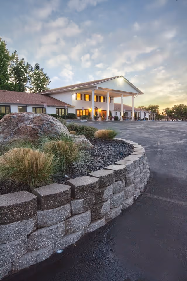 Exterior view of Westview Healthcare Center at dusk, showing a two-story building with large white columns at the entrance, illuminated windows, a curved stone retaining wall with landscaping, and an empty parking lot.