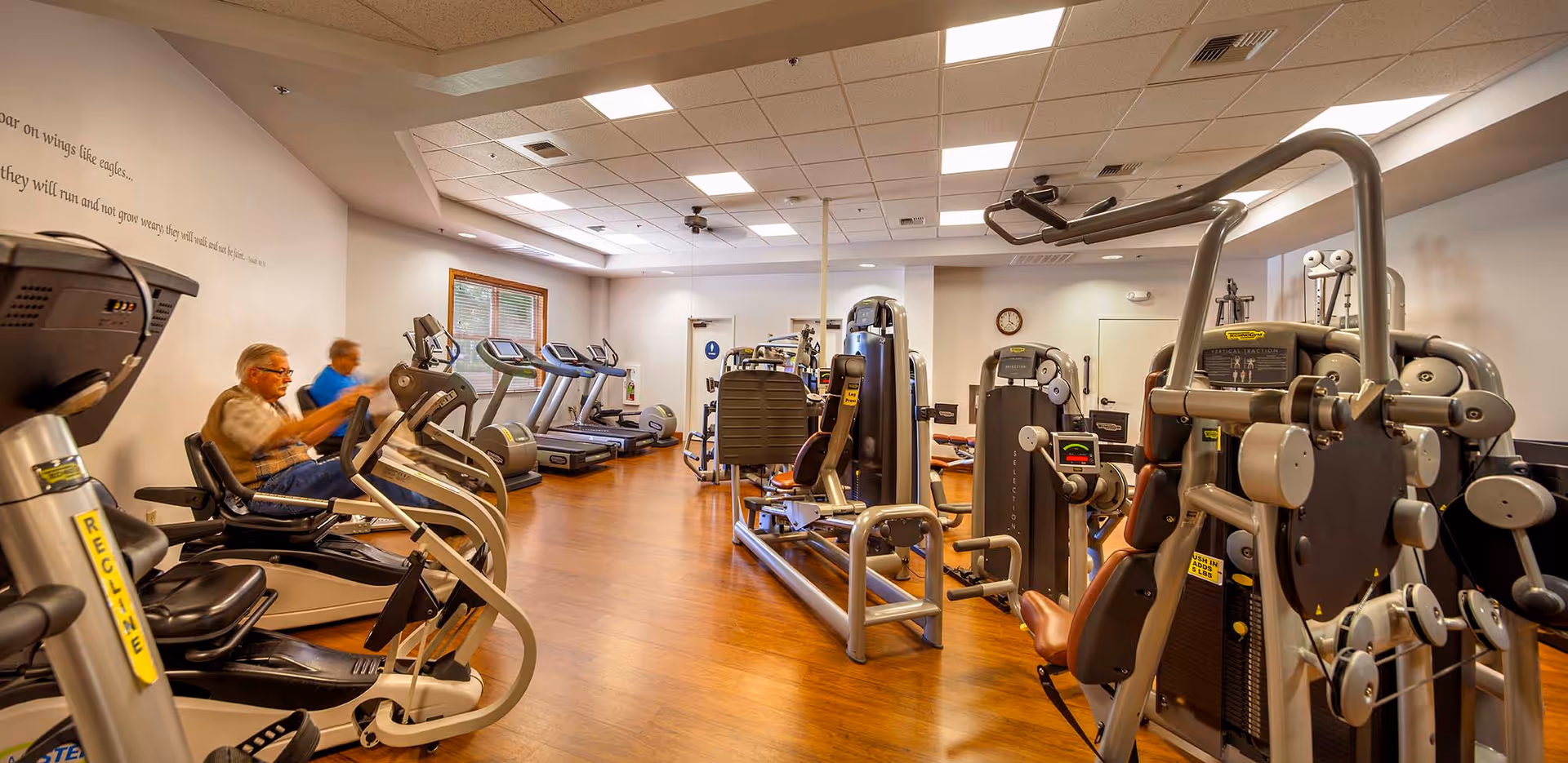 Interior view of a fitness center with various exercise machines including treadmills, stationary bikes, and weight machines. Two elderly individuals are using the equipment. The room has wooden flooring, white walls, a window, and ceiling lights.