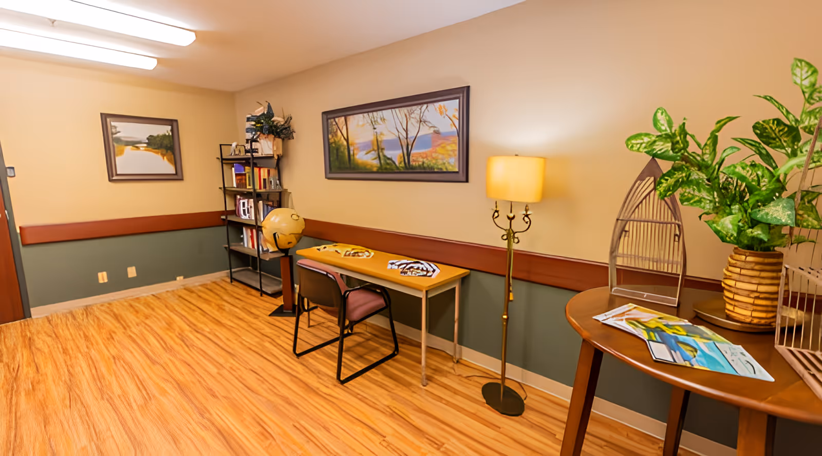 A cozy interior room with wooden flooring and beige walls with a green lower half. The room features a small table with a chair, a standing lamp, a bookshelf with books and decorative items, two framed landscape paintings on the walls, and a round wooden table with a potted plant and some papers on it.