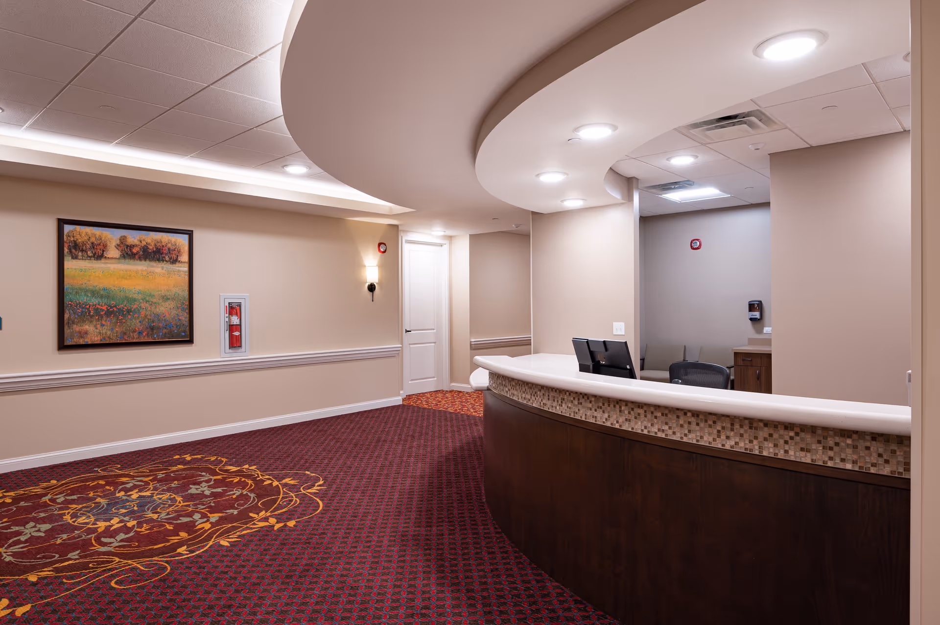 Interior view of a senior living facility reception area with a curved wooden front desk, computer monitors, beige walls, a colorful patterned carpet, a landscape painting on the wall, and ceiling lights.
