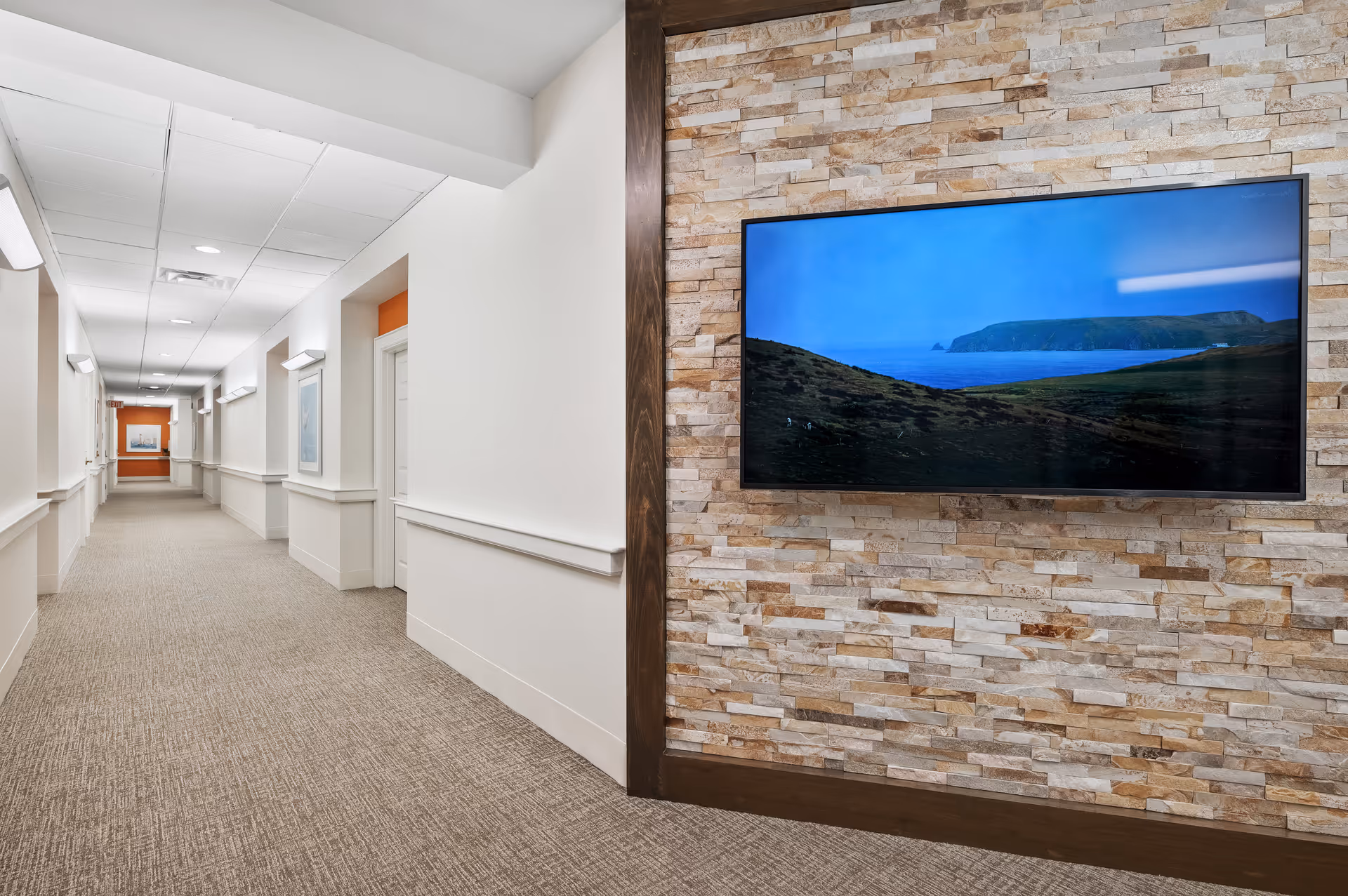 A long, well-lit hallway in a senior living facility with beige carpet and white walls. On the right side, there is a stone accent wall with a large flat-screen TV mounted on it displaying a landscape image of hills and water. Several doors and framed pictures line the hallway.