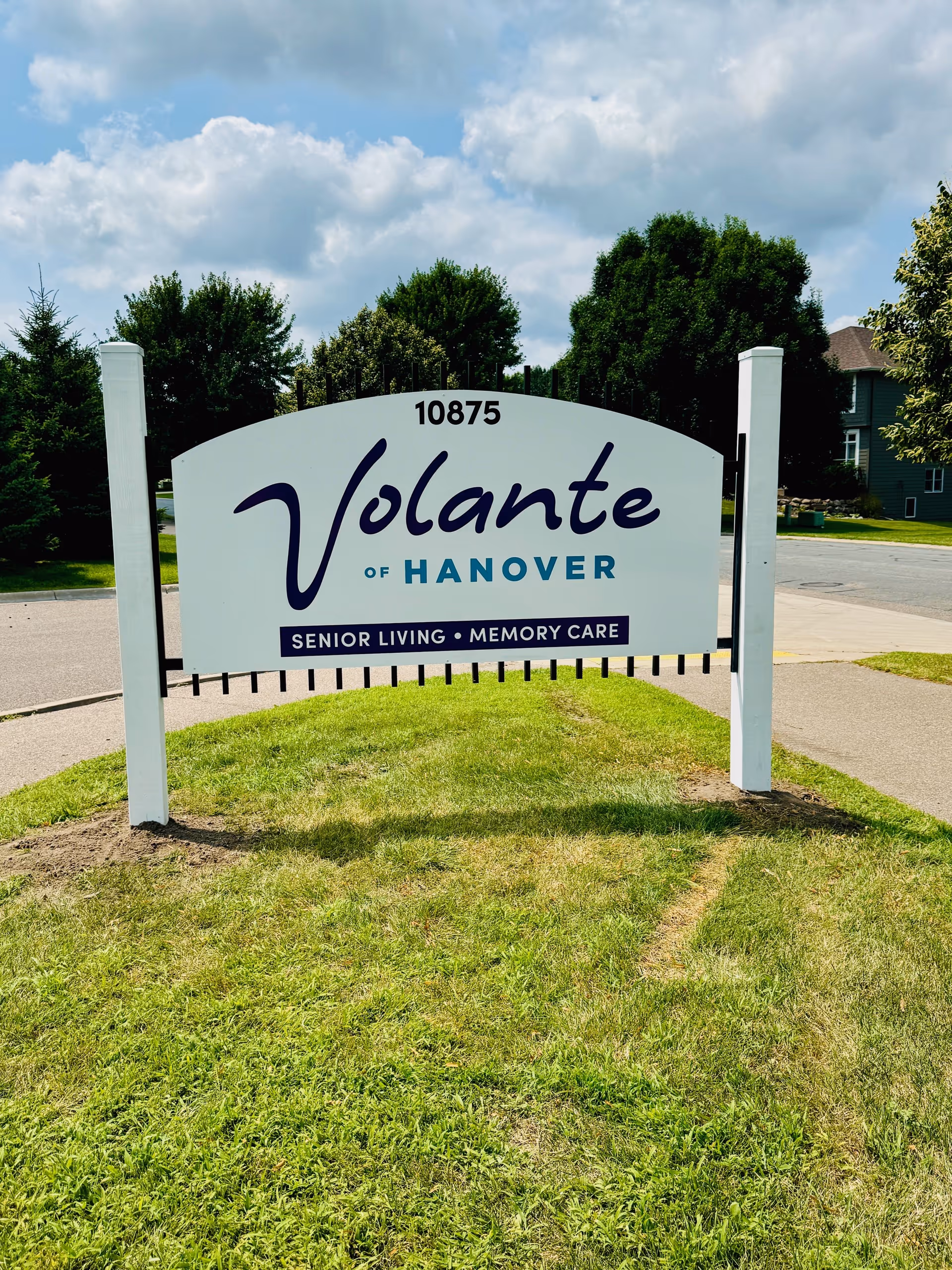 A white sign with black and blue text that reads '10875 Volante of Hanover Senior Living • Memory Care' standing on a grassy area near a sidewalk with trees and houses in the background under a partly cloudy sky.