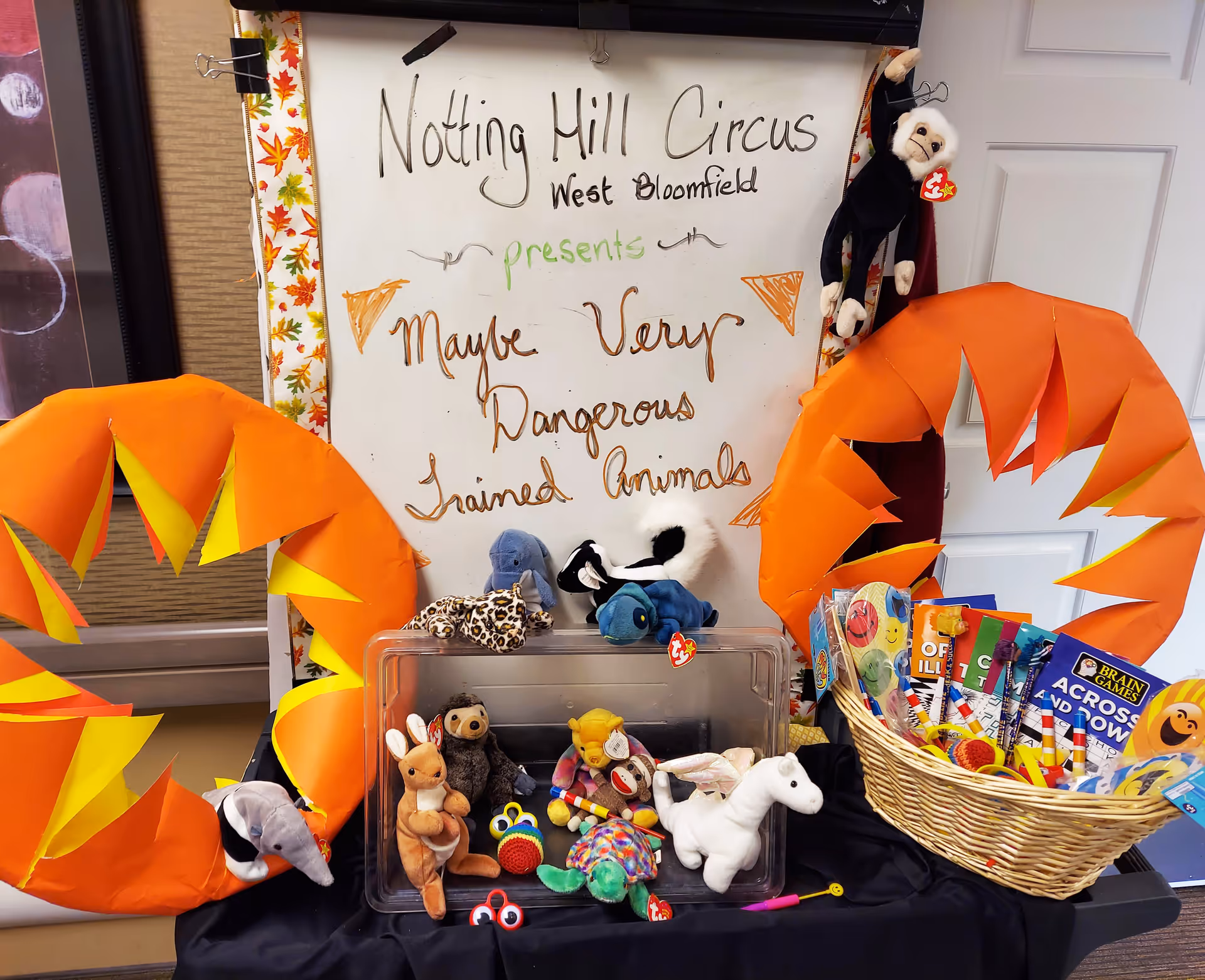 A display board with the text 'Notting Hill Circus West Bloomfield presents Maybe Very Dangerous Trained Animals' surrounded by orange and yellow paper decorations. Below the board are various small stuffed animals and toys, including a monkey hanging on the right side, and a basket filled with activity books and games on the right.