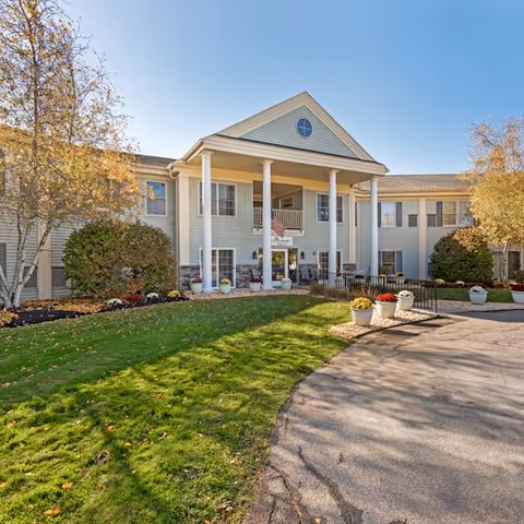 Front exterior view of a two-story senior living facility building with white columns, an American flag hanging near the entrance, landscaped bushes, trees with autumn leaves, and a paved driveway leading to the entrance.
