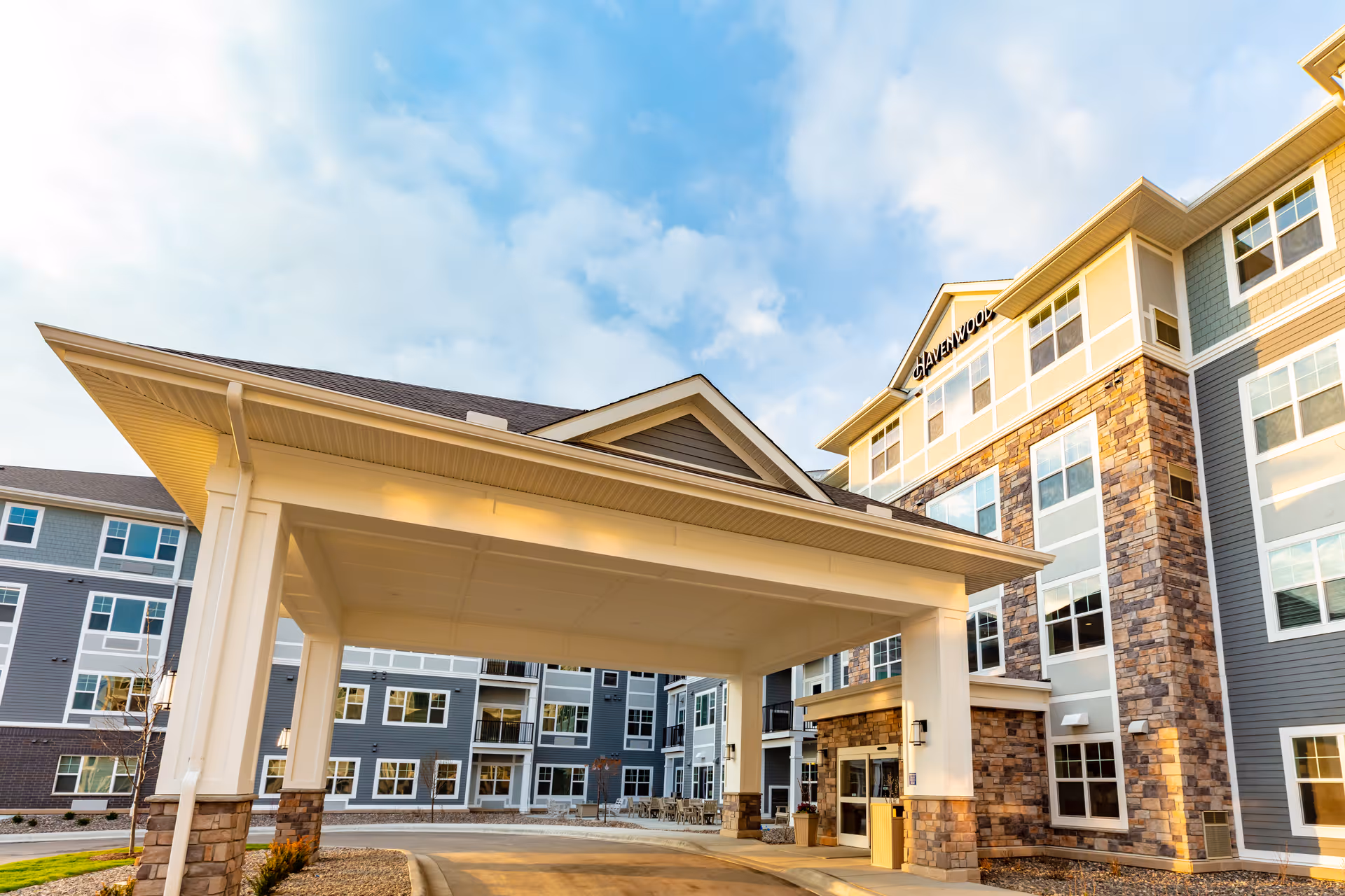 Exterior view of Havenwood of Maple Grove senior living facility showing a large covered entrance with stone and siding facade under a partly cloudy sky.