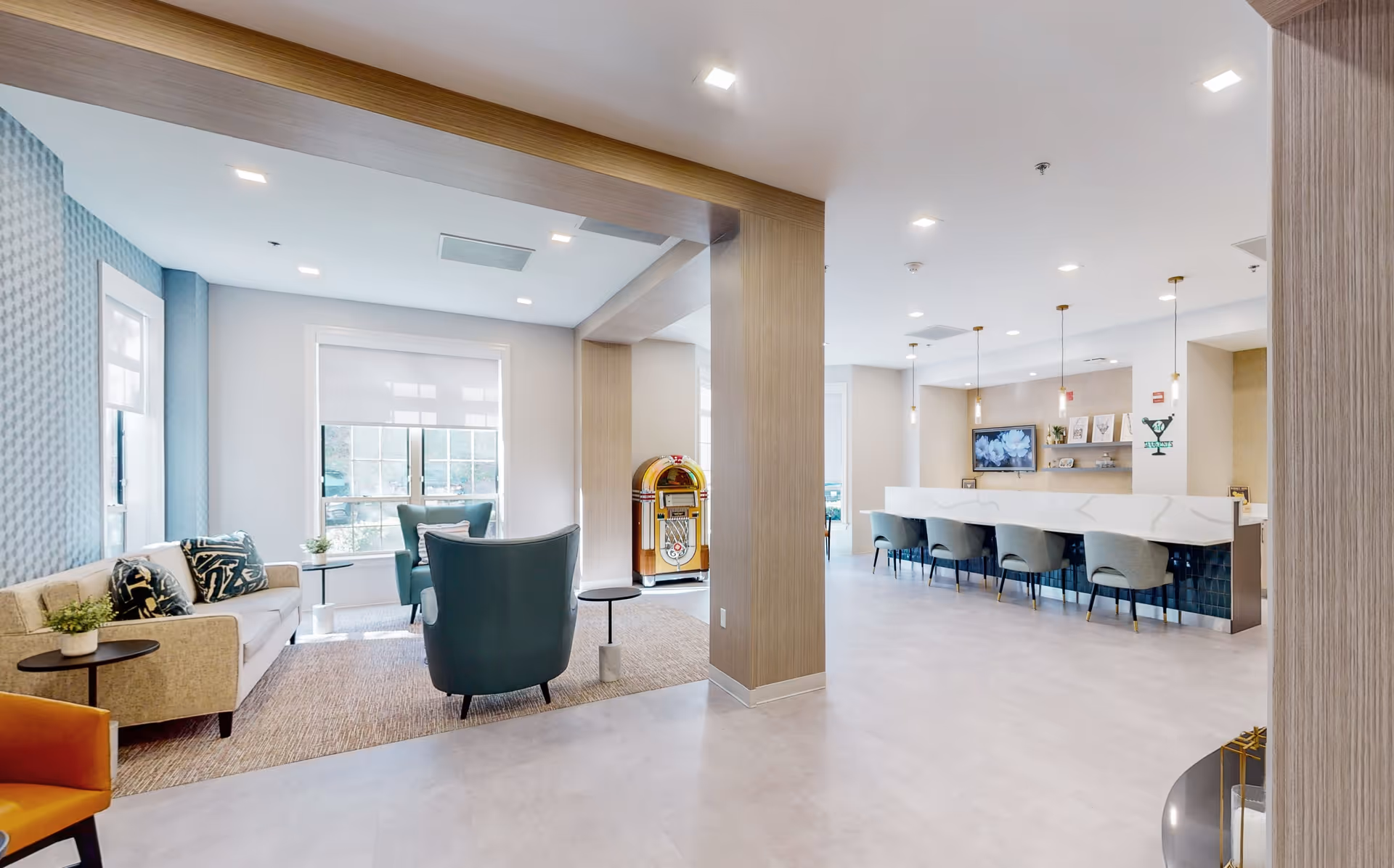 A bright and modern common area in a senior living facility featuring a seating area with a beige sofa, two teal armchairs, and a small round table. There is a jukebox against the wall near large windows with white blinds. To the right, there is a long counter with five teal chairs and pendant lights hanging above. The walls are light-colored with wood accents and a television mounted on the far wall.