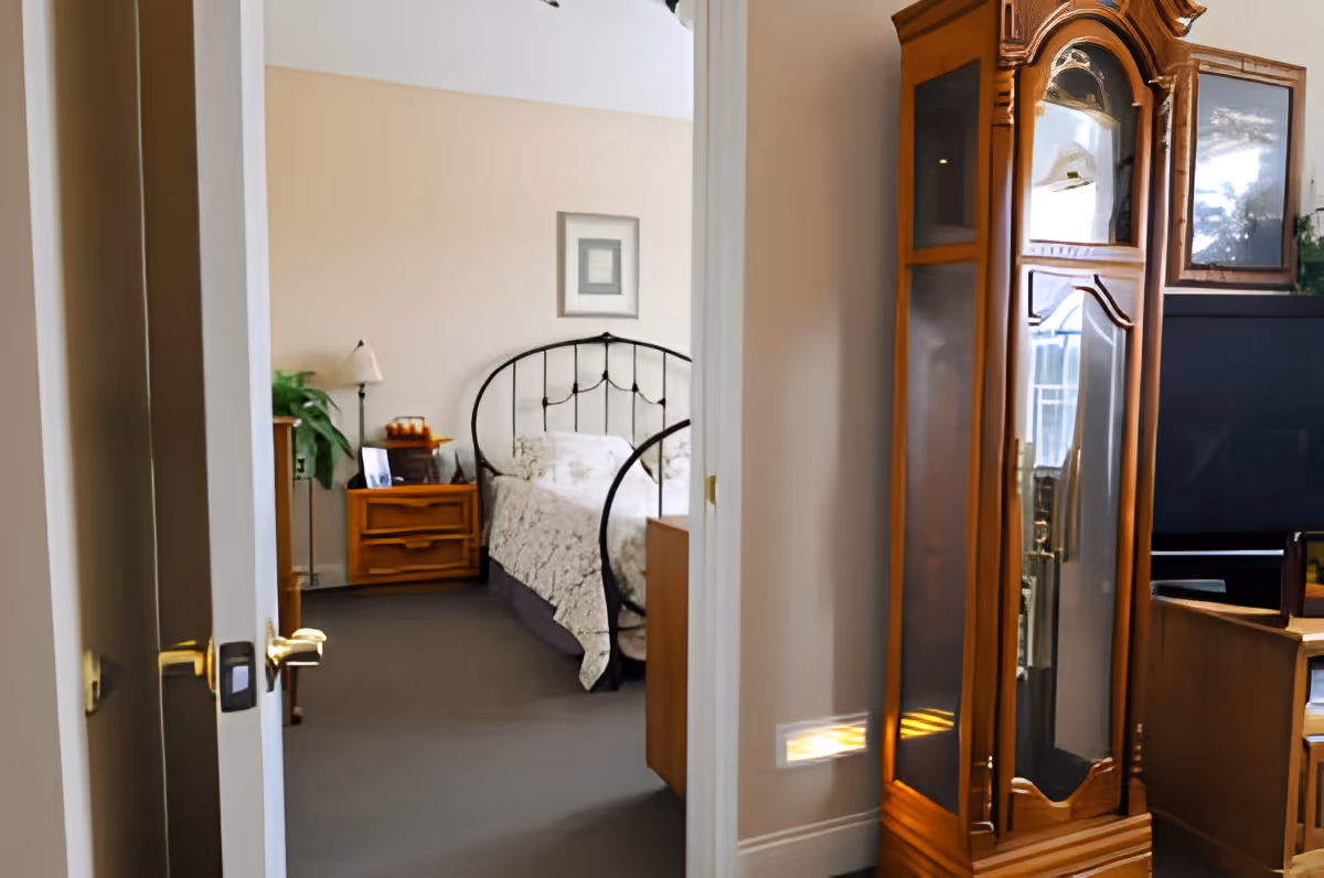 View through an open doorway into a bedroom with a black metal bed frame, floral bedspread, wooden nightstand with a lamp and plant, and a framed picture on the wall. In the foreground, there is a wooden grandfather clock and part of a TV and wooden furniture.