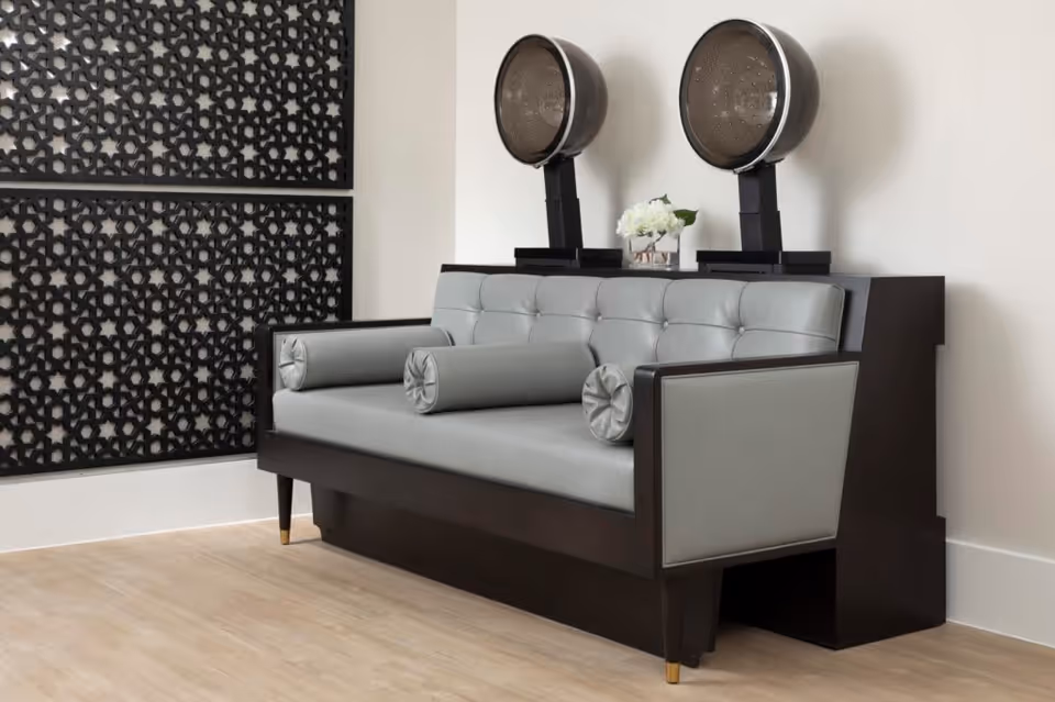 A modern salon area with a gray cushioned bench featuring two cylindrical pillows, positioned in front of two vintage-style hair dryers mounted on a dark wooden stand. The background includes a decorative black lattice panel on the wall and light wood flooring.