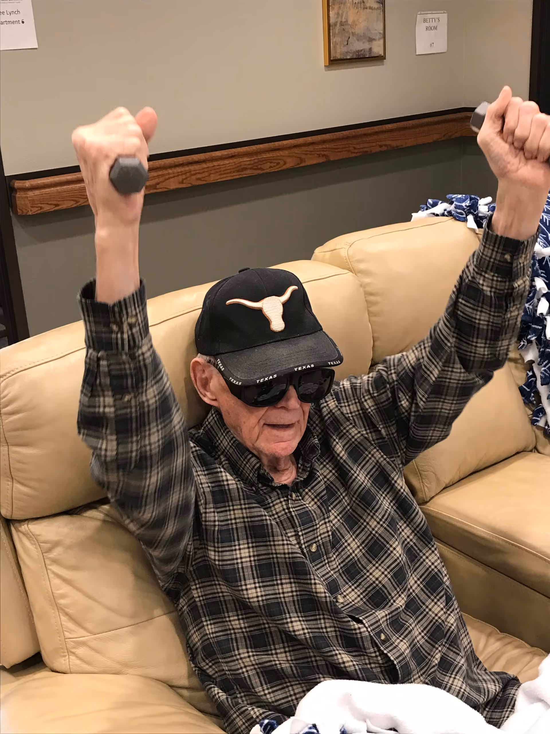 An elderly man wearing a black cap with a longhorn logo and dark sunglasses is sitting on a beige leather couch, lifting small dumbbells with both hands raised above his head. He is dressed in a plaid shirt and appears to be exercising indoors in a room with neutral-colored walls and a wooden handrail.