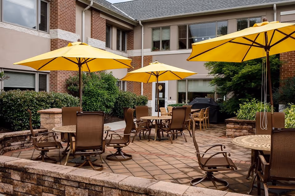Outdoor patio area at Phoebe Berks with multiple round tables surrounded by brown chairs and shaded by large yellow umbrellas. The patio is paved with tiles and bordered by low stone walls, with greenery and bushes around the perimeter. The building exterior is visible in the background.
