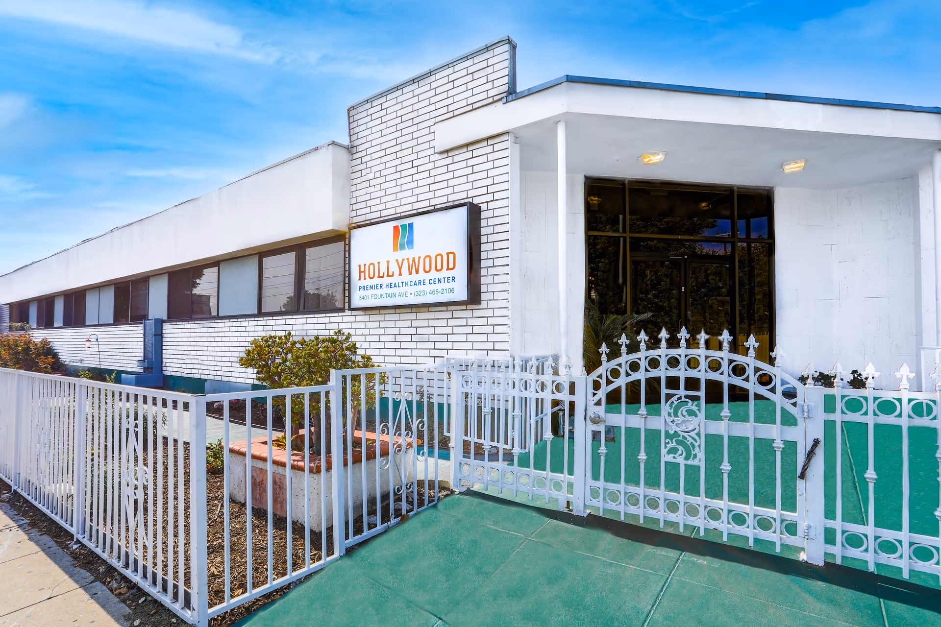 Exterior view of Hollywood Premier Healthcare Center showing a white brick building with a white metal fence and gate in front. The entrance has a covered porch area with glass doors and the facility's sign is mounted on the wall near the entrance.