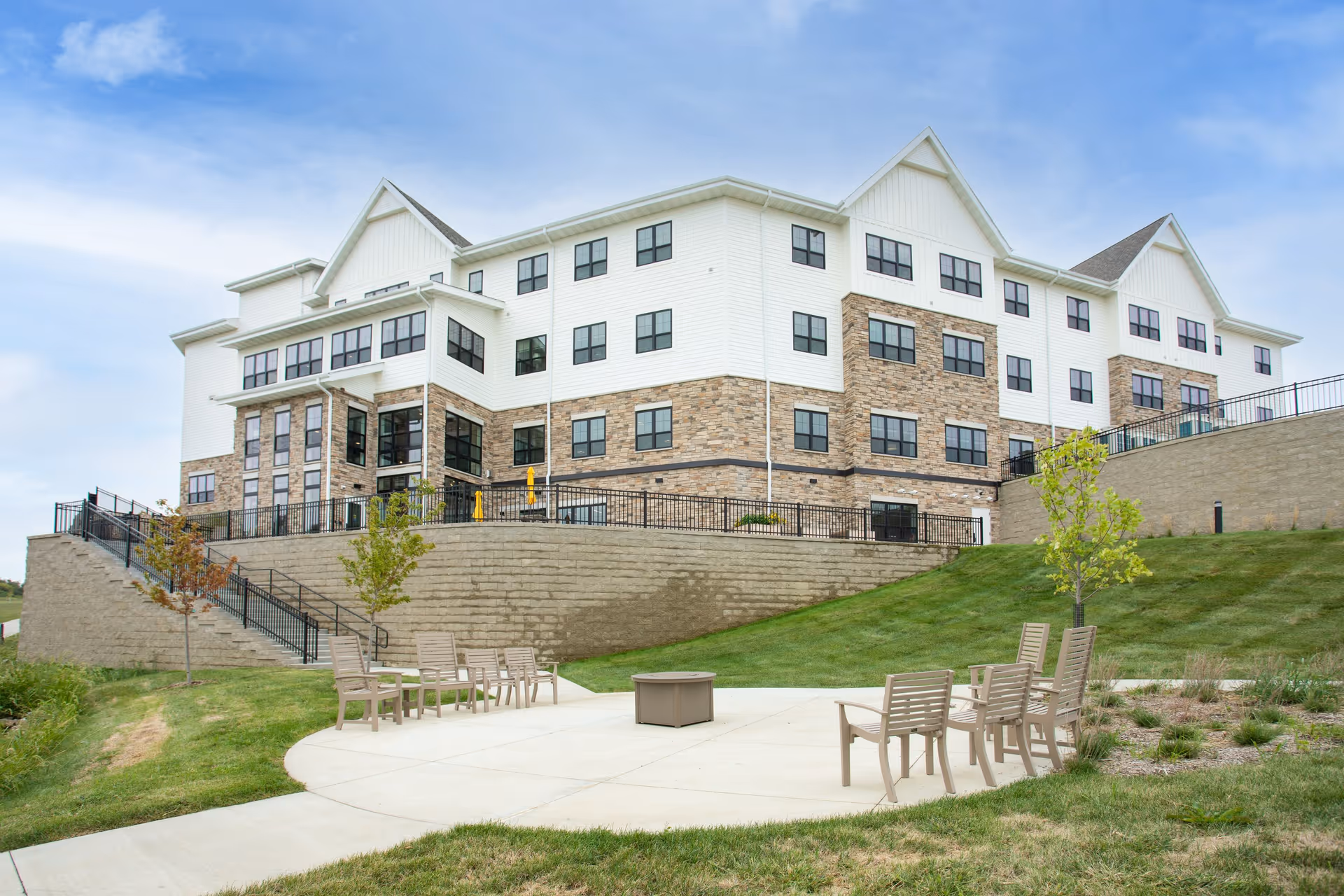 Exterior view of a multi-story senior living facility building with white siding and stone accents. In the foreground, there is a circular concrete patio area with several beige chairs arranged around a central fire pit. The area is surrounded by green grass and small trees under a partly cloudy blue sky.