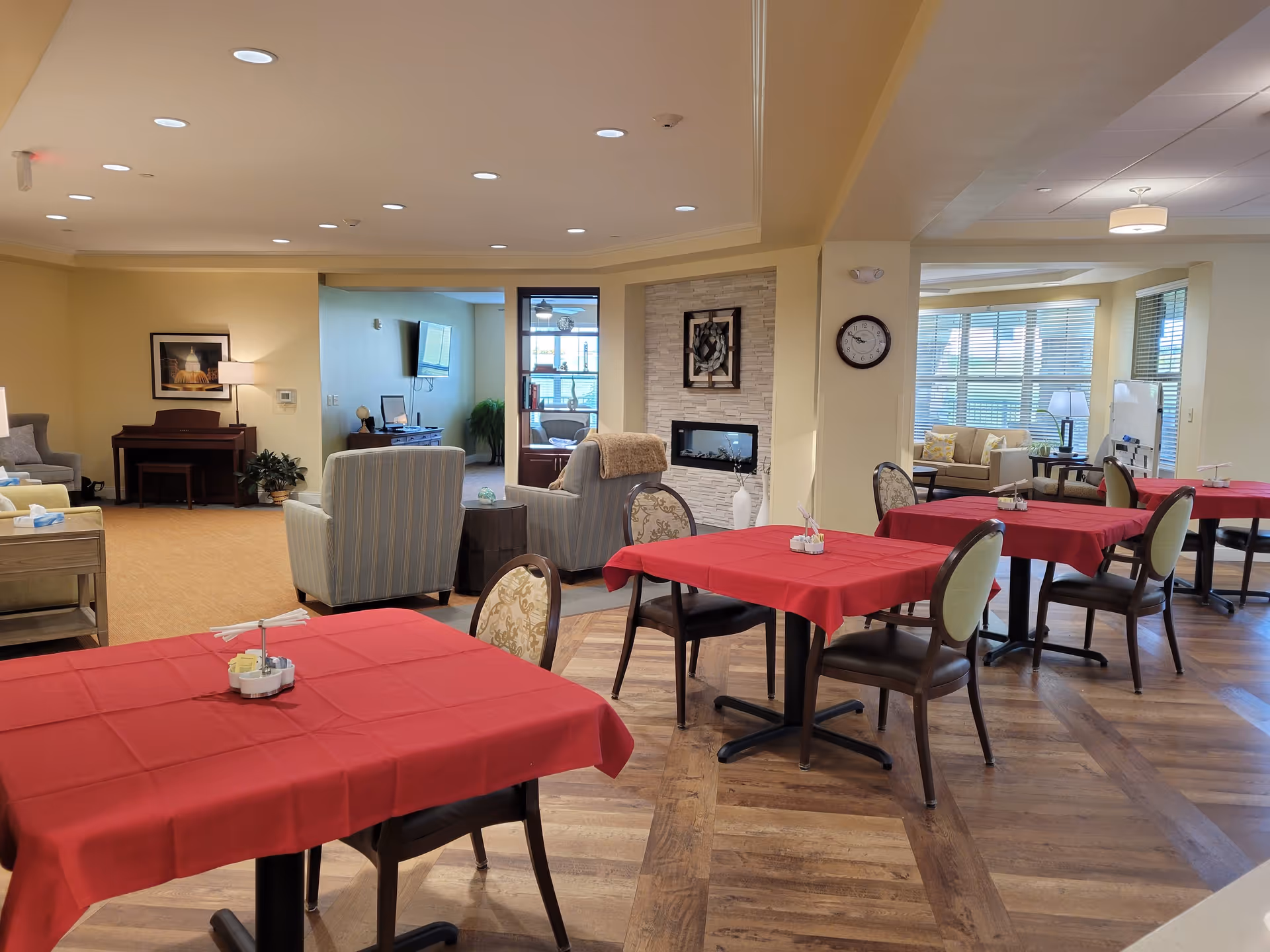 Interior view of a senior living facility dining area with tables covered in red tablecloths and chairs around them. In the background, there is a cozy sitting area with armchairs, a fireplace, a piano, and a wall clock. Large windows allow natural light to brighten the space.
