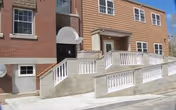 Exterior view of a multi-story building with a brick and siding facade. There is a wheelchair-accessible ramp with white railings leading to a door with a small awning above it. Windows are visible on both floors, and the sky is clear and blue.