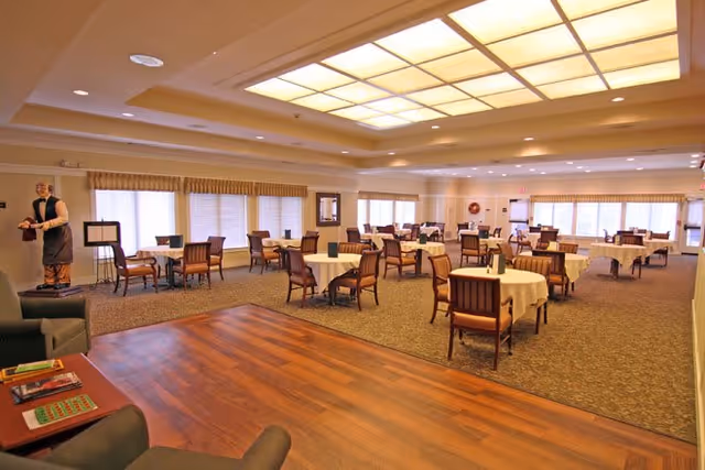 A spacious dining room with multiple round tables covered with white tablecloths, each surrounded by wooden chairs. The room has large windows with blinds and valances, a wooden floor section in the foreground, and a ceiling with large recessed lighting panels. A statue of a waiter holding a tray is visible near the left side of the room.