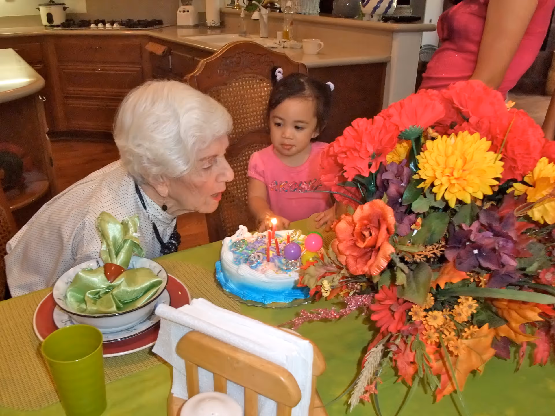 An elderly woman and a young girl leaning over a small birthday cake with lit candles on a table decorated with a large bouquet of flowers.