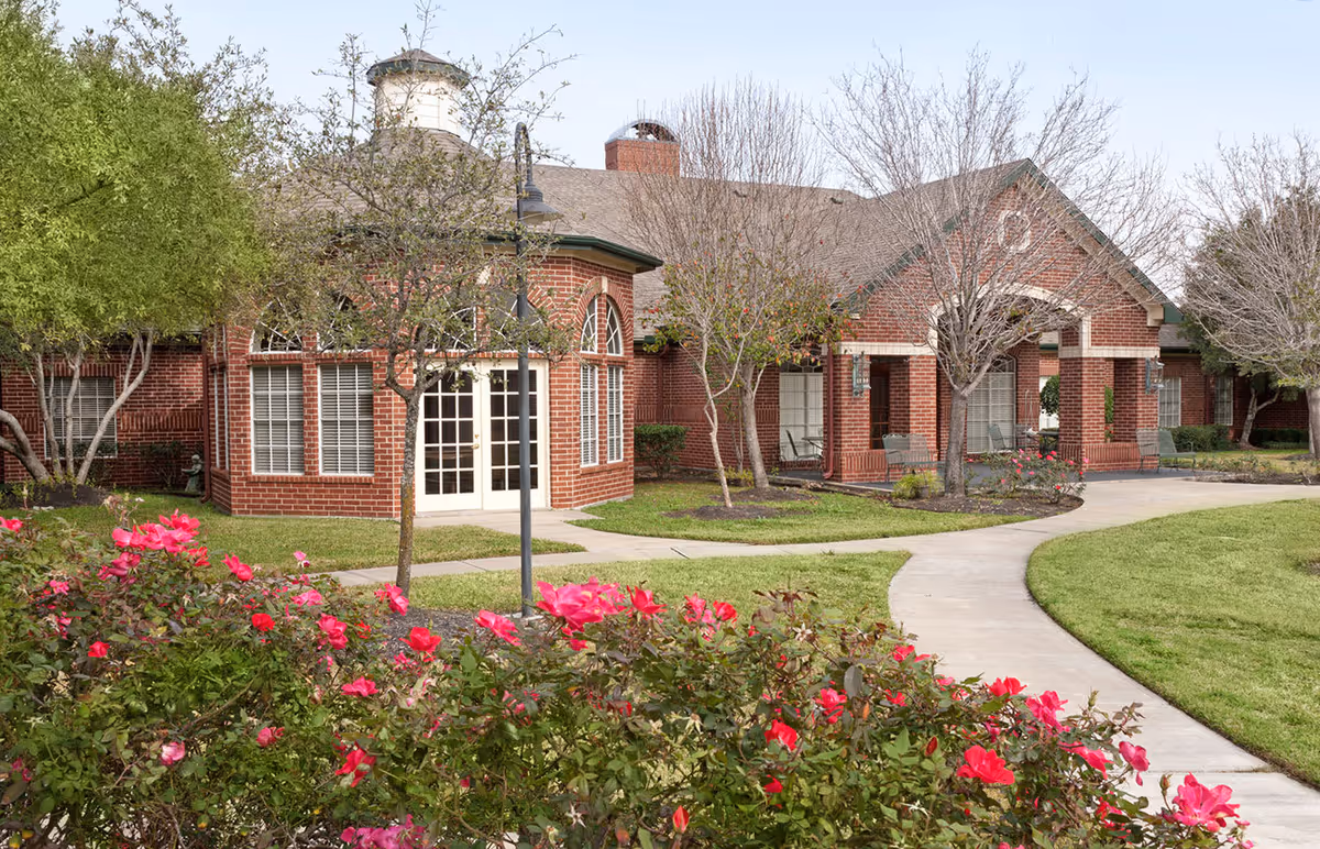 Exterior view of a brick building with large windows and a white door, surrounded by a garden with blooming pink flowers, green grass, and trees. A curved concrete pathway leads to the entrance of the building.