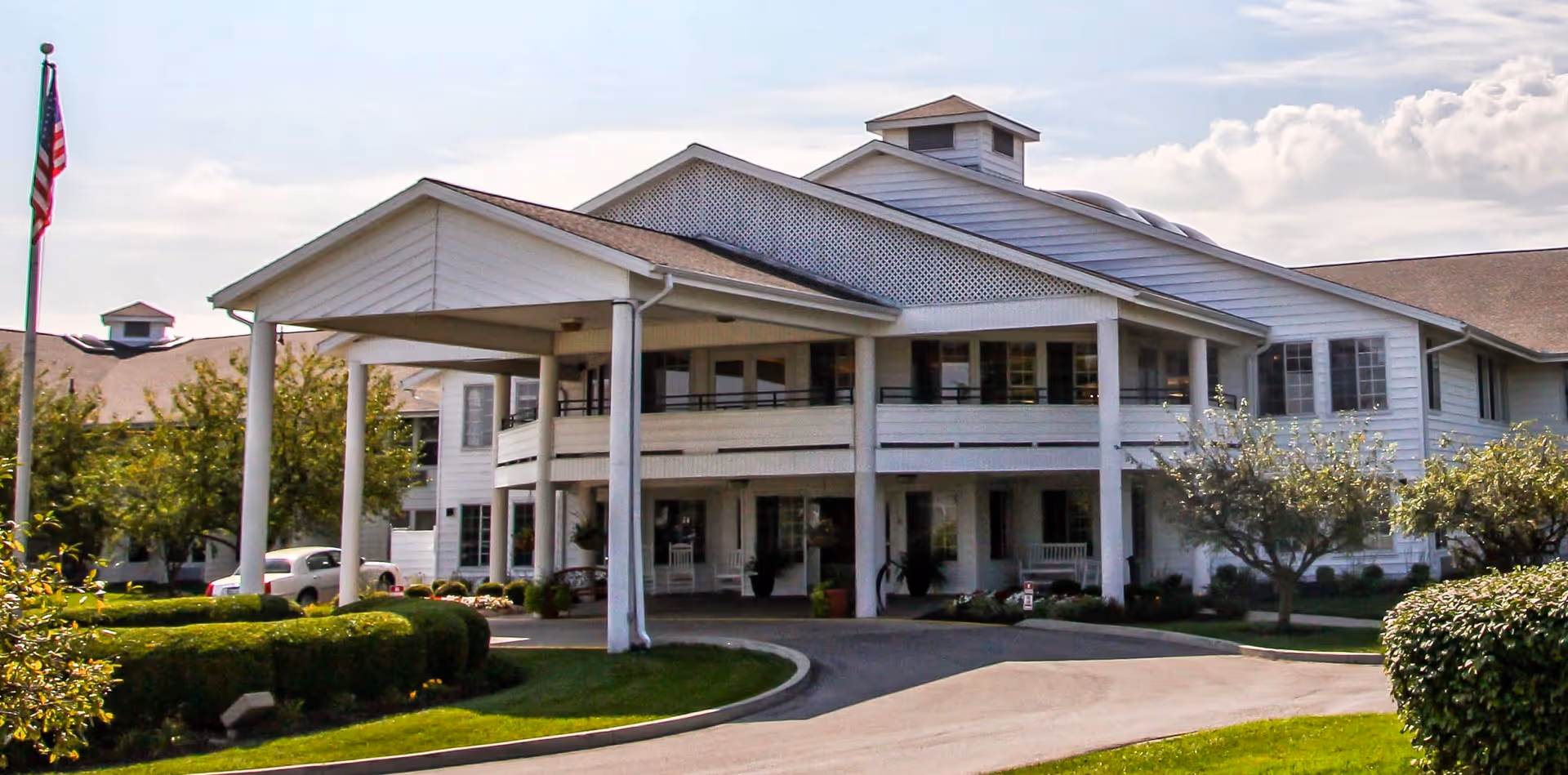 Front exterior view of a large white senior living facility building with a covered entrance supported by columns, surrounded by well-maintained landscaping including bushes, trees, and a lawn. An American flag is visible on a flagpole to the left.
