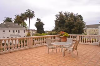 Outdoor terrace with terracotta tiled floor, a round glass table with four chairs, a potted plant on the table, and a balustrade railing. Trees and buildings are visible in the background under a cloudy sky.