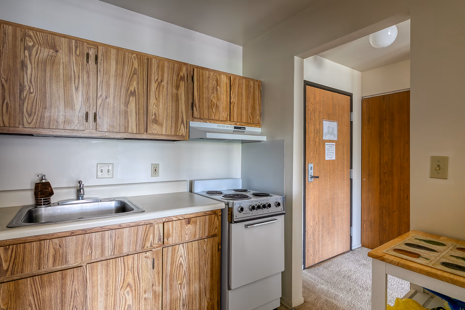 Small kitchen area with wood grain cabinets, a stainless steel sink with a soap dispenser, a white electric stove with four burners, and a wooden door in the background. There is a small table with a tiled surface on the right side.