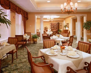 Elegantly furnished dining room with multiple tables set for a meal, upholstered chairs, chandeliers, and decorative columns.