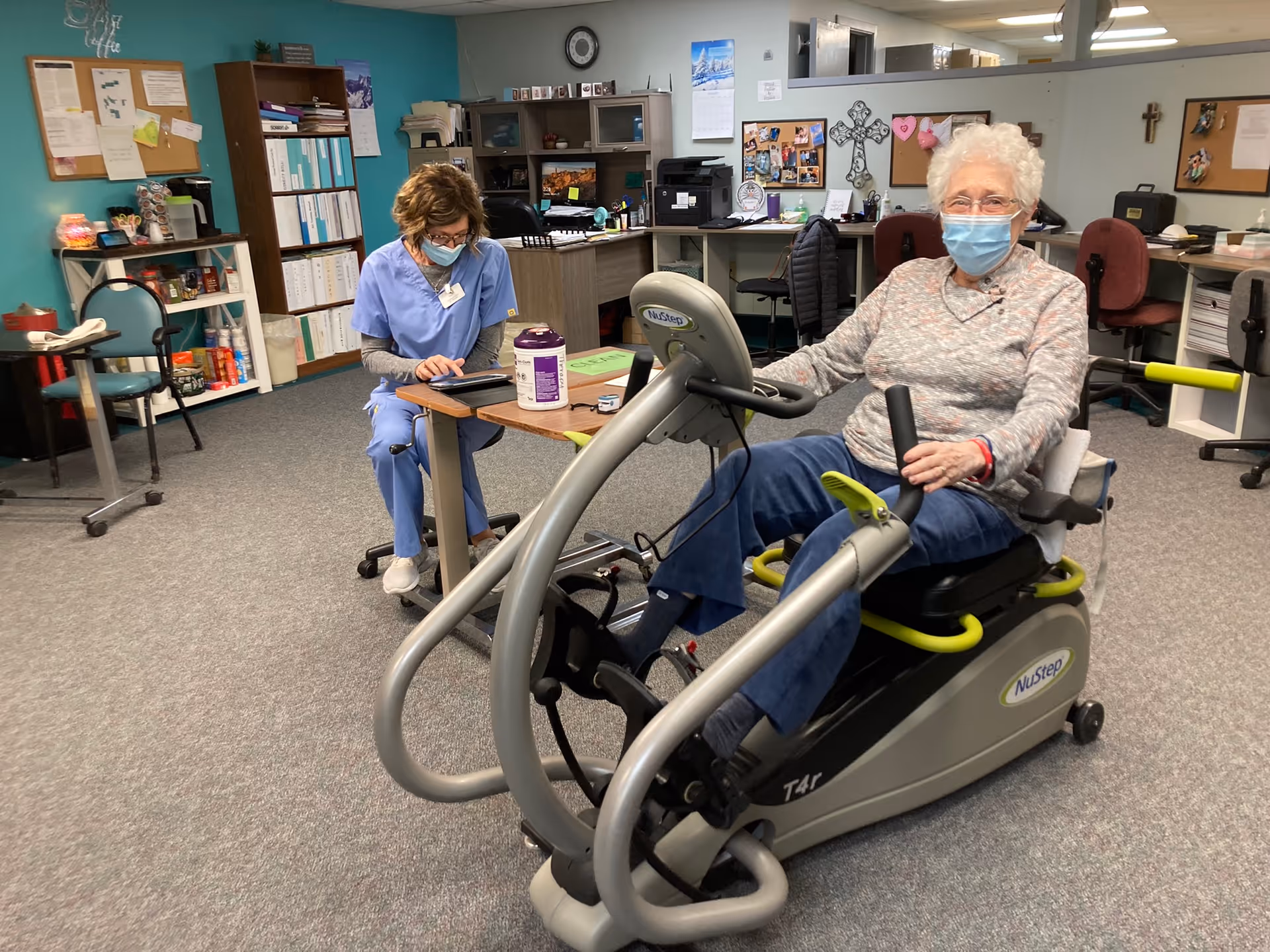 An elderly woman wearing a mask is exercising on a NuStep recumbent cross trainer machine in an indoor room. A healthcare worker in blue scrubs and a mask is sitting at a desk nearby, looking at a tablet. The room has office furniture, shelves with binders, and bulletin boards on the walls.