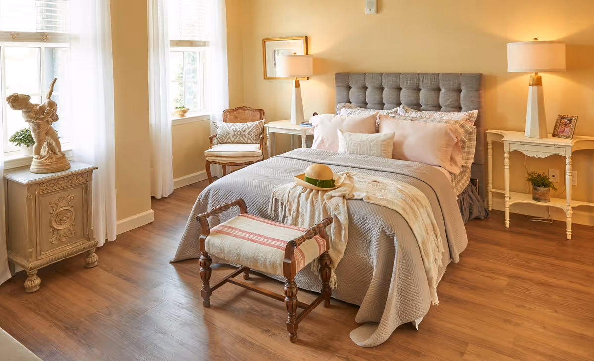 A cozy bedroom with a neatly made bed featuring a gray quilted headboard, multiple pillows, and a beige throw blanket with a hat on top. The room has wooden flooring, two windows with white curtains, a wooden chair with a cushion, two white bedside tables each with a lamp, a small framed picture, and decorative plants. There is also a small upholstered bench at the foot of the bed and a decorative statue on a small ornate cabinet near the window.